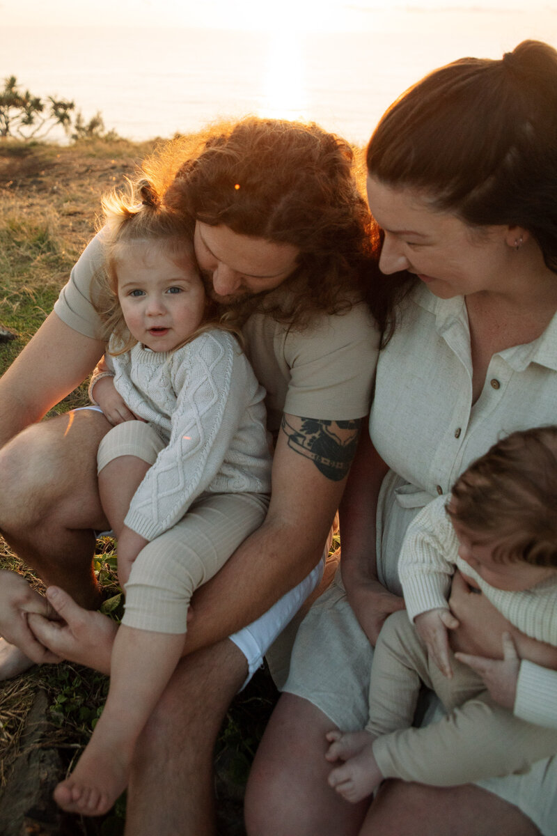 A family of four are cuddled on the cabarita beach headland, the photois taken at sunrise they are cosy and playful with their baby and toddler. The sunrise light is gently lighting their hair and skin. 