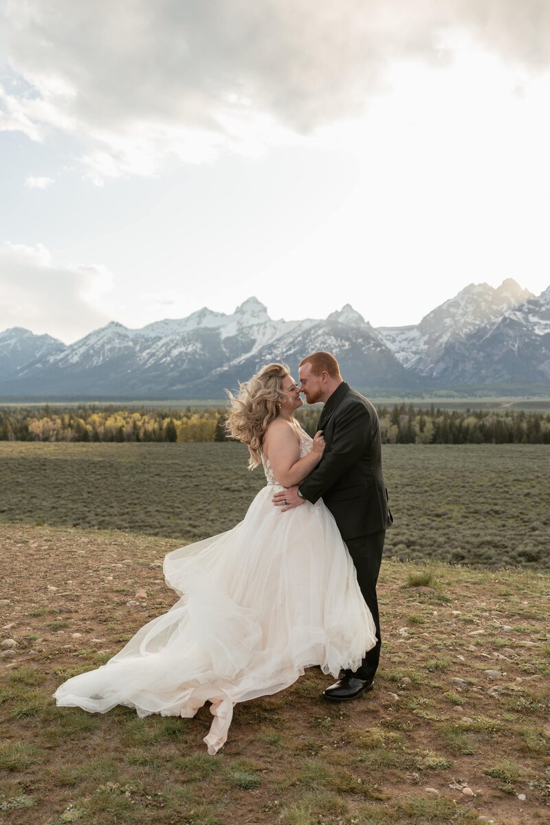 A couple shares an intimate moment during their sunset elopement in Grand Teton National Park, with snow-capped mountains in the background and the bride’s dress flowing in the wind, captured by Sydney Breann Photography.