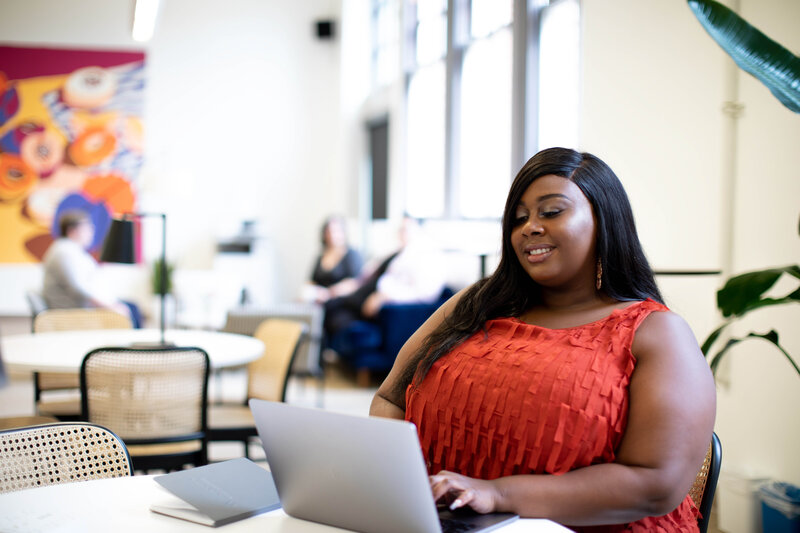 This image shows a plus size, feminine-presenting person of color sitting at a table with a laptop and notebook. They are typing on their laptop, while smiling.