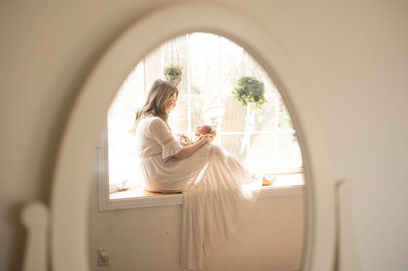 A woman dressed in white sits on a window sill, holding an infant. The scene is reflected in an oval mirror, reminiscent of the sophisticated aesthetics often utilized in Showit web design for luxury photographers. The large window behind them lets in soft, natural light.