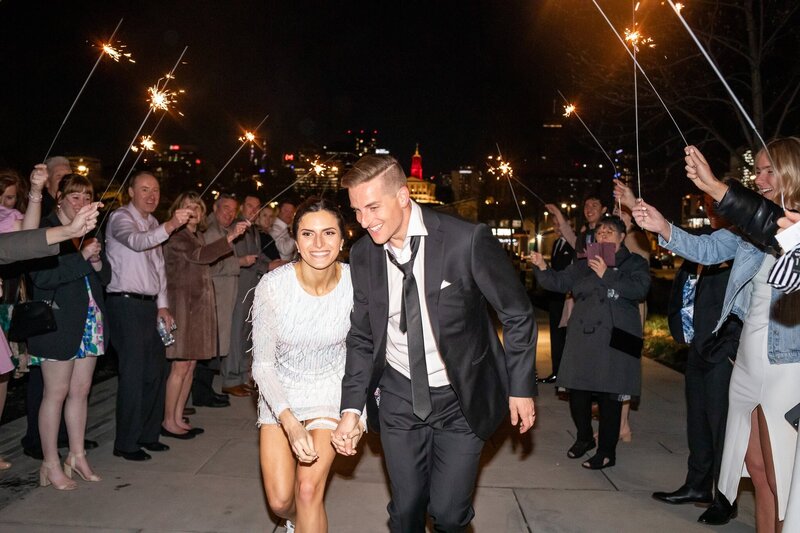 A couple walks through a sparkler exit surrounded by cheering guests, with a glowing skyline twinkling behind them. The bride wears a short sequin dress and beams beside her partner in a dark suit and loosened tie. The energy is electric, the night alive with celebration—perfectly capturing the joy of a city wedding under the lights.