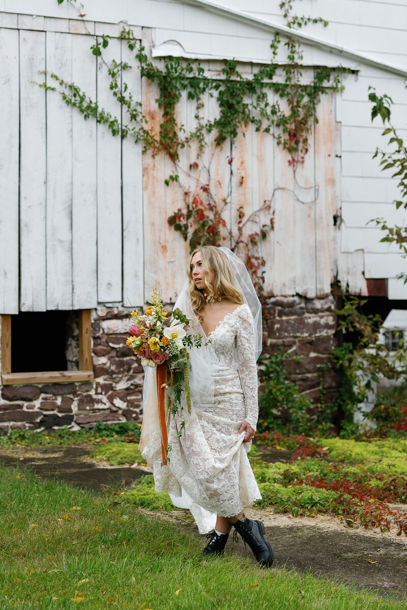 A bride stands outside in boots, lifting her skirts in front of a stone and wood panel barn