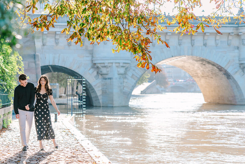 Man and woman walking hand in hand by the Seine river, smiling during a romantic Paris proposal photoshoot near Pont Neuf on Île de la Cité.