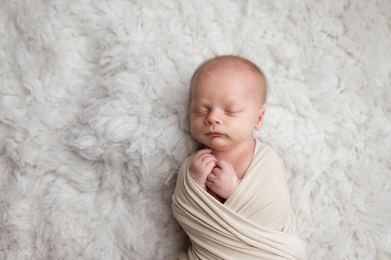 newborn wrapped in a tan blanket while on a soft rug