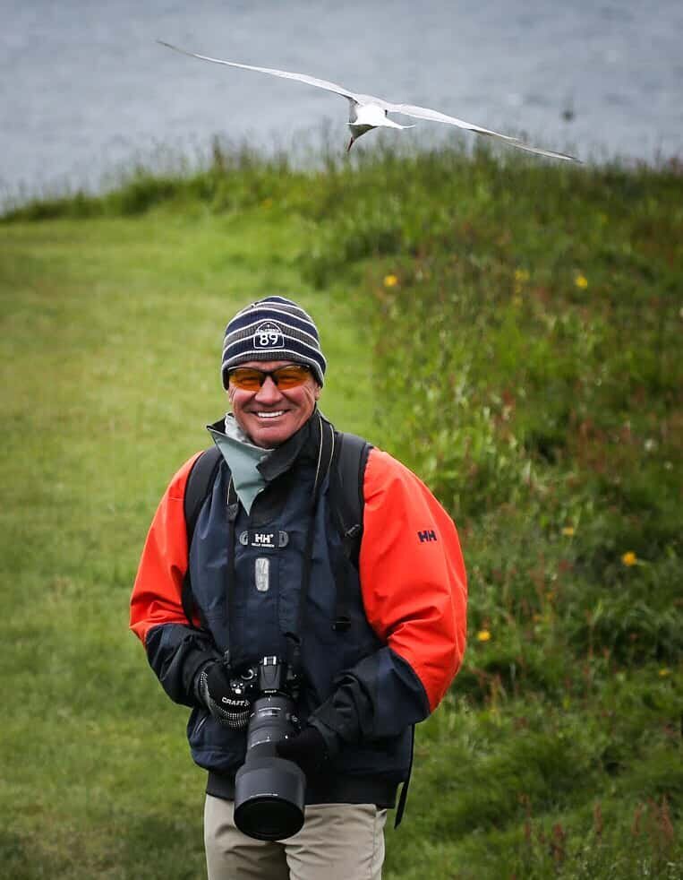 Photographer smiling and dressed in a blue and orange rain jacket holding a very large camera under an Icelandic bird that is swooping down on his head