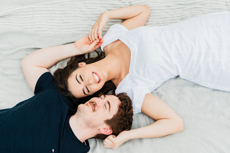 Wedding photographer in Dallas captures a happy couple lying down on a blanket outside, both smiling and happy, she is in a plain white dress and their eyes are closed. a great couples pose for engagement photos.
