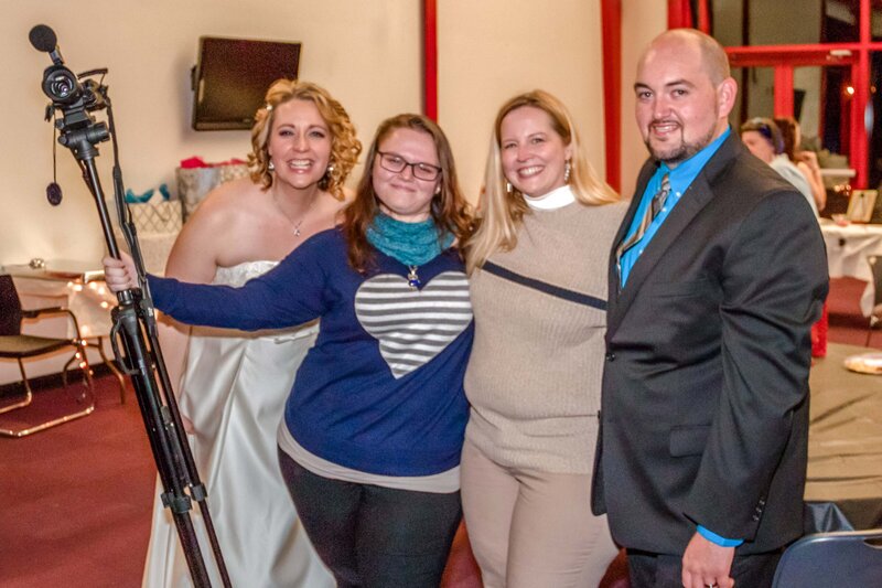 Behind-the-scenes photo of a smiling bride and groom posing with the wedding photography and videography team, including a woman holding a video camera tripod.