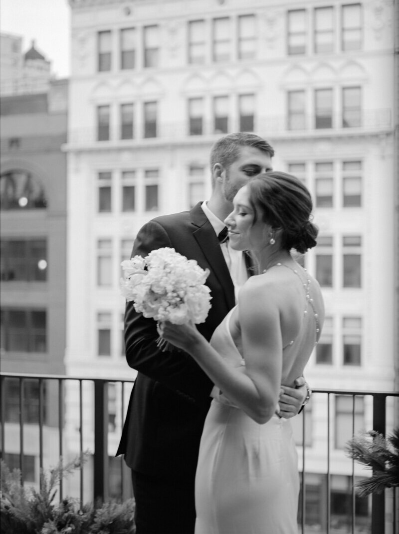 bride in backless wedding dress with pearl straps smiles, kissed by her husband on Shinola Hotel Detroit balcony