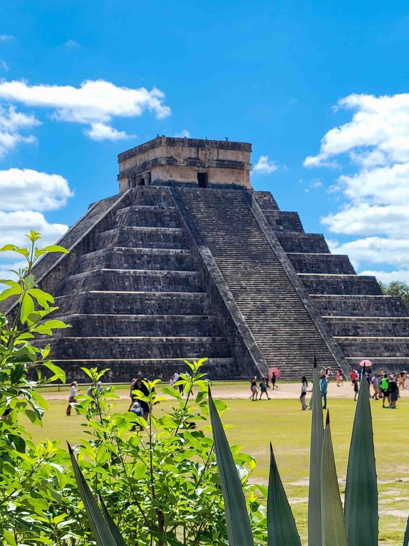 El Castillo pyramid at Chichén Itzá in Yucatán, Mexico, rising above a green landscape, framed by spiky agave plants in the foreground under a bright blue sky ©Stephanie Dosch | theViatrix Custom Mexico Travel Plan