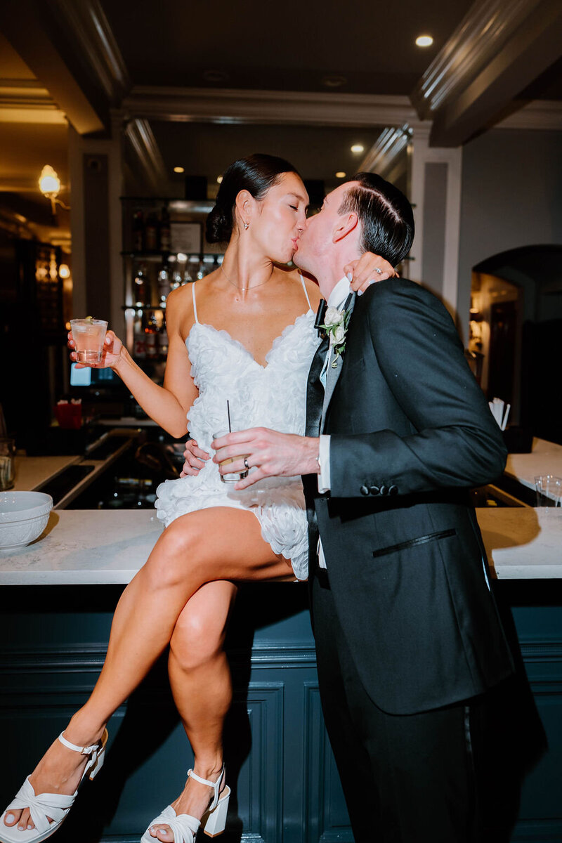 Rhode Island Wedding Photographers | A couple in wedding attire shares a kiss at a bar. The bride, in a short lace dress, sits on the counter holding a drink. 