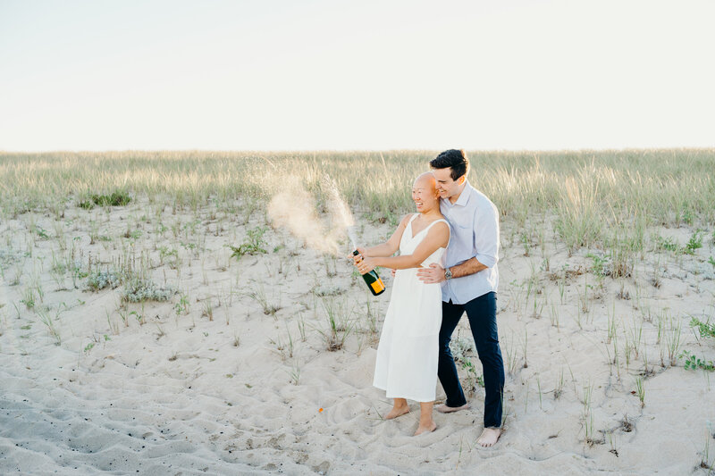 engaged couple popping champagne at sunset on chatham lighthouse beach in massachusetts