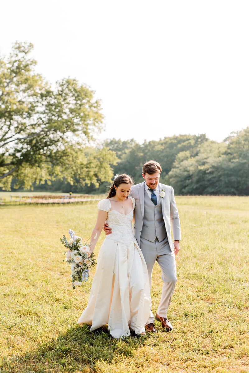 bride and groom walking hand-in-hand across a sunny meadow, the bride holding a bouquet of pale flowers