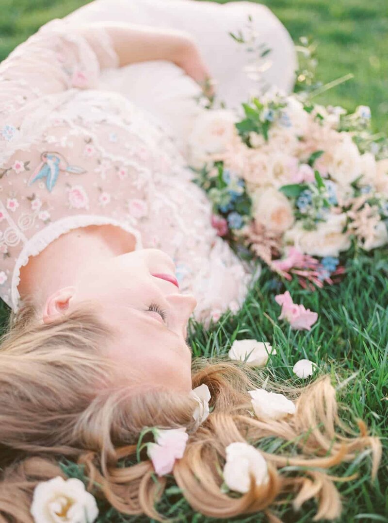 A pregnant woman lays on the ground surrounded by flowers with her blonde hair flowing behind her and she has one hand below her belly