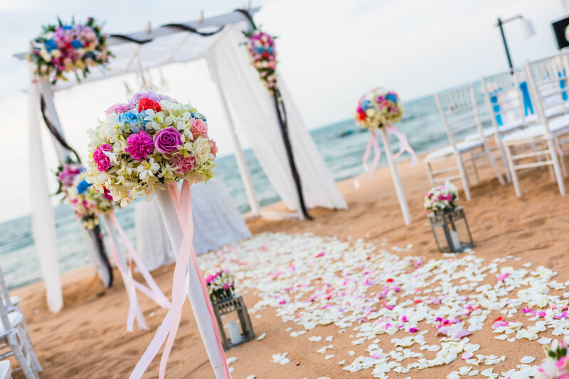 Colorful beach wedding setup with an arch draped in white fabric, floral arrangements in pink, purple, and blue, and a petal-covered aisle leading toward the ocean.