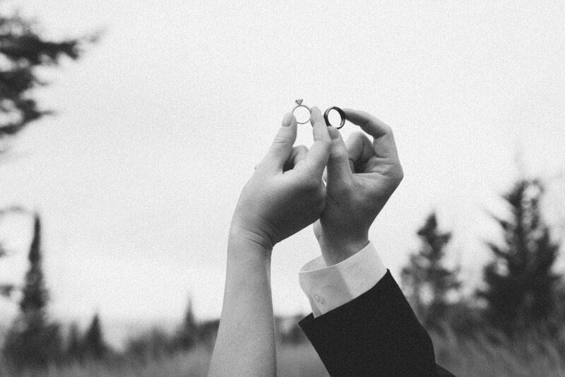Couple eloping in the forest holds their wedding rings in the air, surrounded by towering pine trees. 