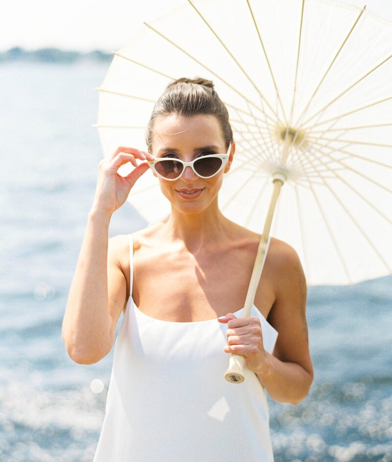 Rhode Island Wedding Photographer | Woman in sunglasses holds a white parasol by the ocean. She wears a white dress, smiling softly, conveying a sunny, relaxed vibe with a clear blue sea backdrop.