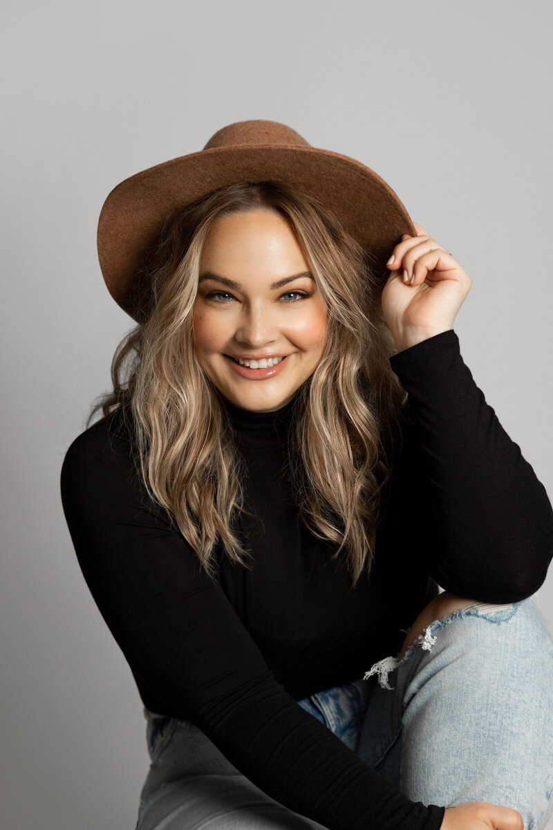 Premium studio headshots of woman in black turtleneck and ripped jeans. Sitting on the ground, she is smiling at the camera and touching a tan felt hat. She has long blonde curly hair and smiling at the camera. Image was captured against a white backdrop in top London, Ontario headshot studio by Ogg Photography.