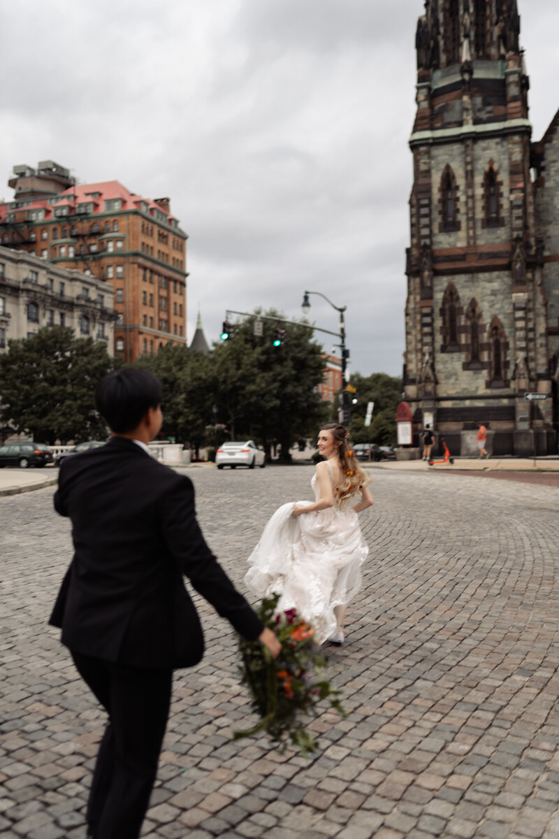 A bride running away jokingly from her groom through a city the groom is holding her bouquet reaching out to her.