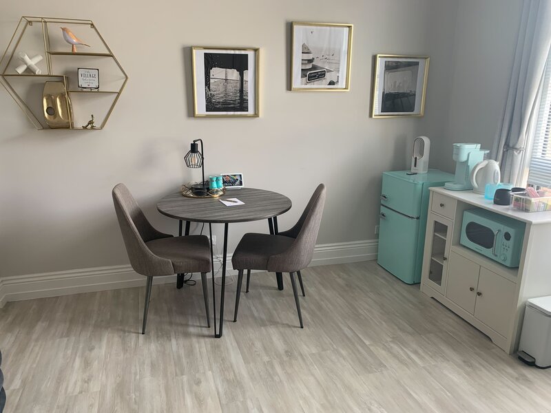 A picture of a suite's dining area. A round table with two armless chairs. Against the wall is a mini-fridge and a cabinet holding a microwave, coffee maker, electric kettle, and a box of coffees and teas. The microwave and fridge are in a vintage style and mint-green. There is art on the walls - black and white photographs - and a shelf with small figurines.