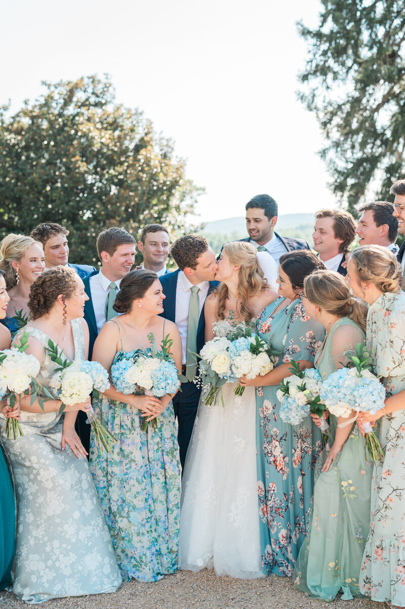 A newlywed couple kissing with their wedding parties cheering around them