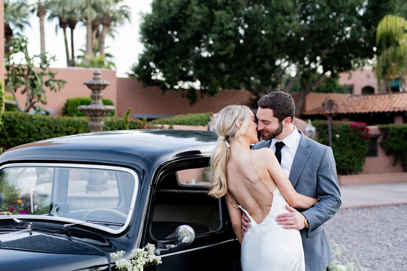 Couple in wedding attire kissing next to a vintage car.
