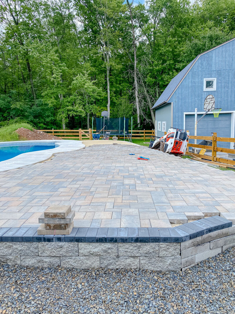 A paver patio pool deck nearly finished with a pool on one side and a blue barn or garage on the other side