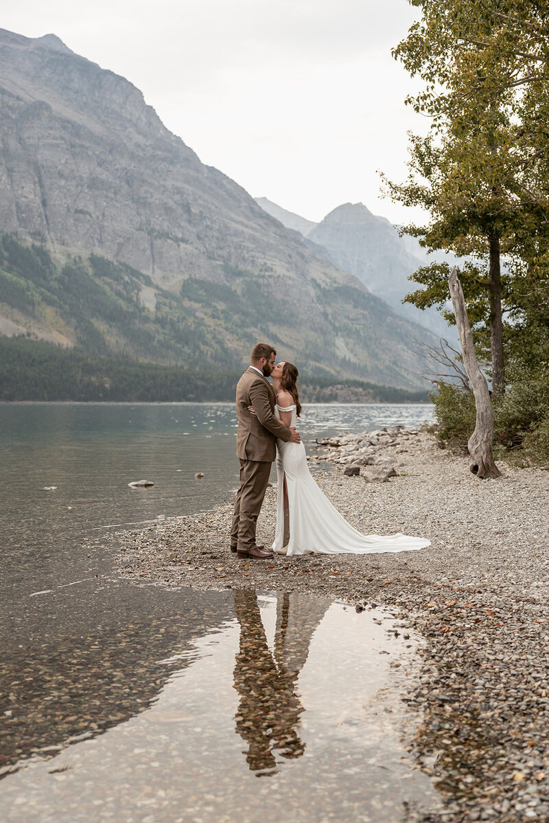 Bride and groom share a kiss on a rocky lakeshore surrounded by mountain views and calm water reflections.