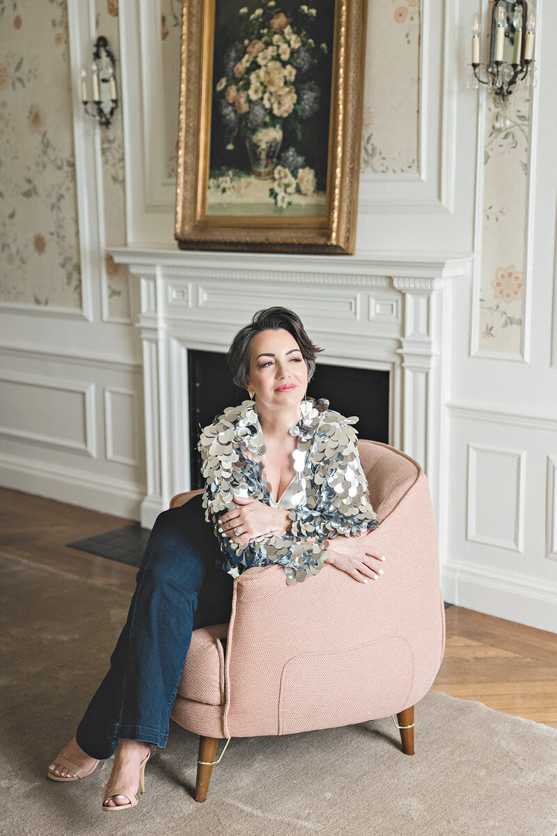 Editorial-style portrait of Kari Dawson seated in a soft pink chair wearing a floral statement top, exuding calm authority and elegant confidence as a Detroit-based wedding photographer who honors quiet moments