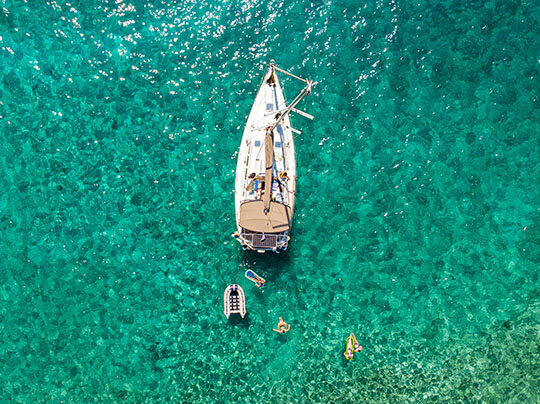 Segelyacht von oben auf dem Meer in tükisblauen Wasser und vier Menschen auf Luftmatratzen im Wasser