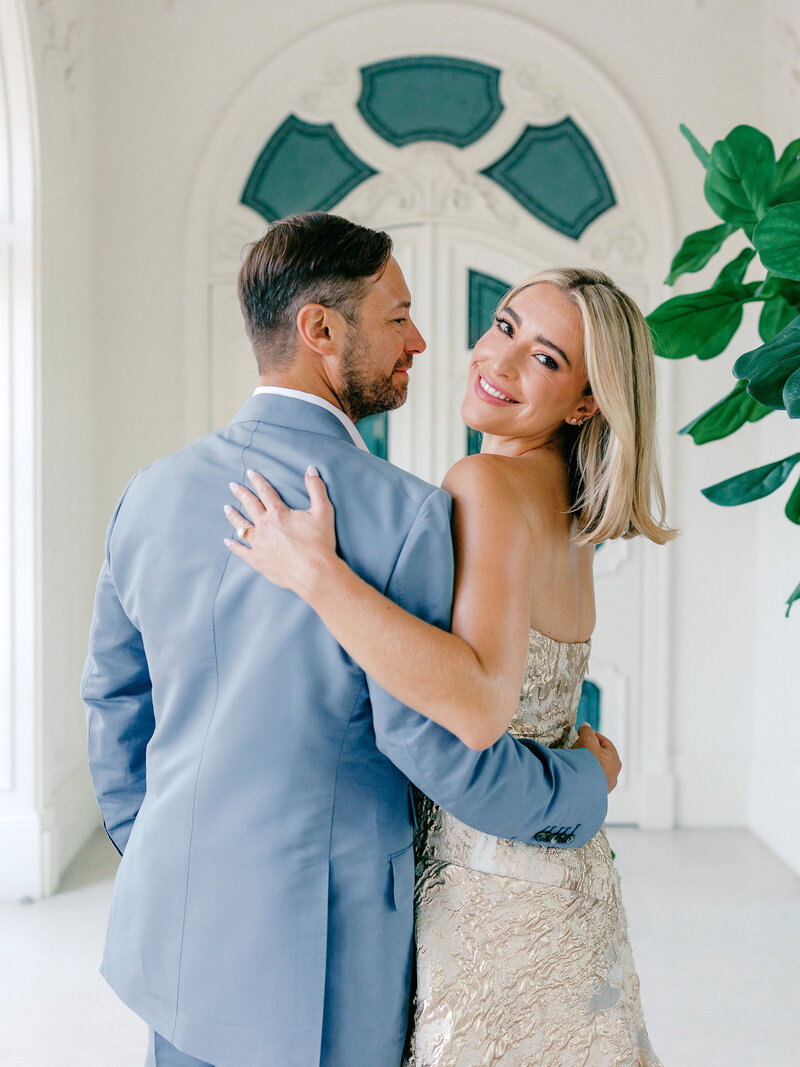 Bride in gold wedding dress looks over shoulder toward the camera while groom in a light blue suit looks at her