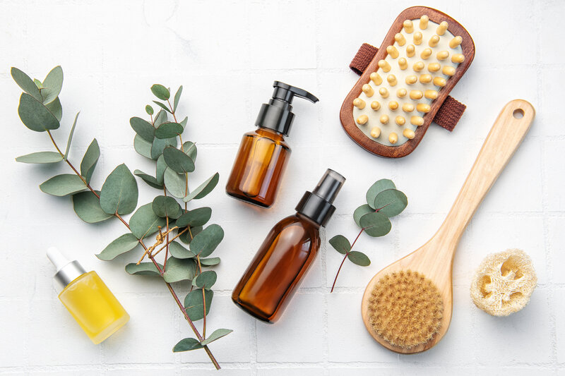 Flat lay of body care tools including amber bottles, dry brush, and eucalyptus leaves on a white background