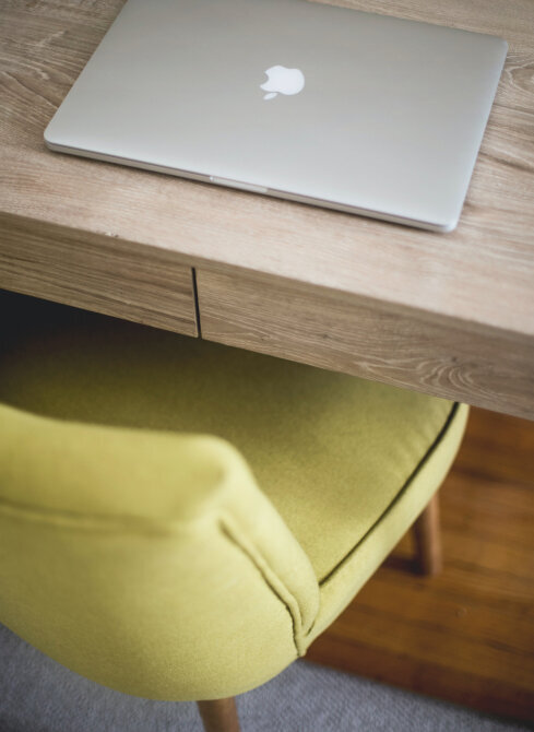 This is a photo of a modern workspace featuring a closed silver laptop, possibly a MacBook, resting on a wooden desk. Below the desk, there is a chair with a yellow-green fabric cover, which complements the warm tones of the wood. The focus on the laptop and the blurred chair suggests a setting that is ready for work or study. The natural materials and soft color palette give the space a comfortable and inviting feel.