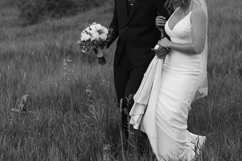 Black and white photo of bride and groom walking through tall grass, holding bouquet, during a domestic wedding.