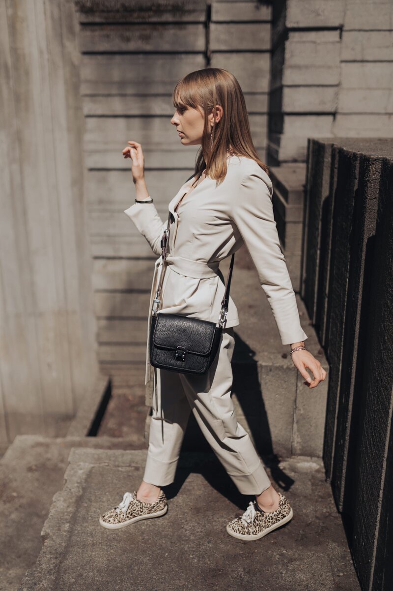 Woman with brown hair turns to the side wearing a khaki jumpsuit and cheetah sneakers showcasing her small, black purse.