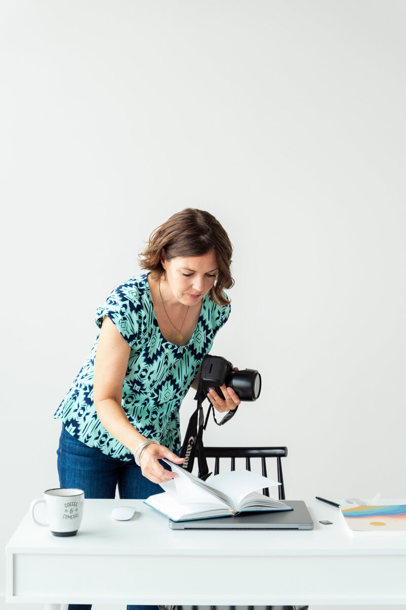 Calgary brand storytelling photographer, Kate Daniel, a woman with wavy brown hair, wearing a patterned blue and green blouse, stands at a white desk while holding a DSLR camera in one hand and flipping through a notebook with the other. The desk is neatly arranged with a laptop, a coffee mug that reads Coffee & Cameras, a mouse, and a notebook with colourful pages. The background is a clean, minimalist white wall.