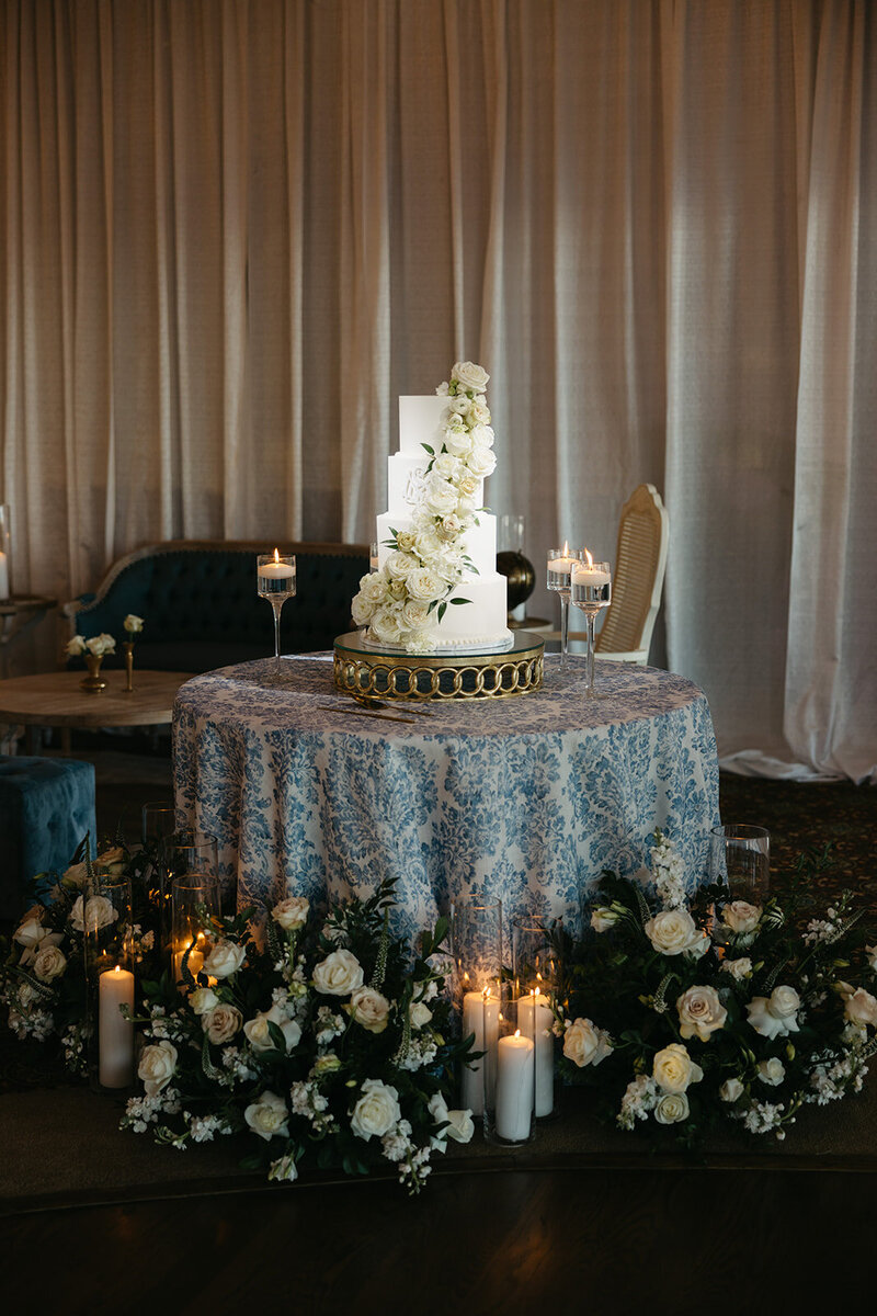 Grand white wedding cake adorned with roses, surrounded by florals and candlelight, styled by The Eventful Gals for a romantic reception.
