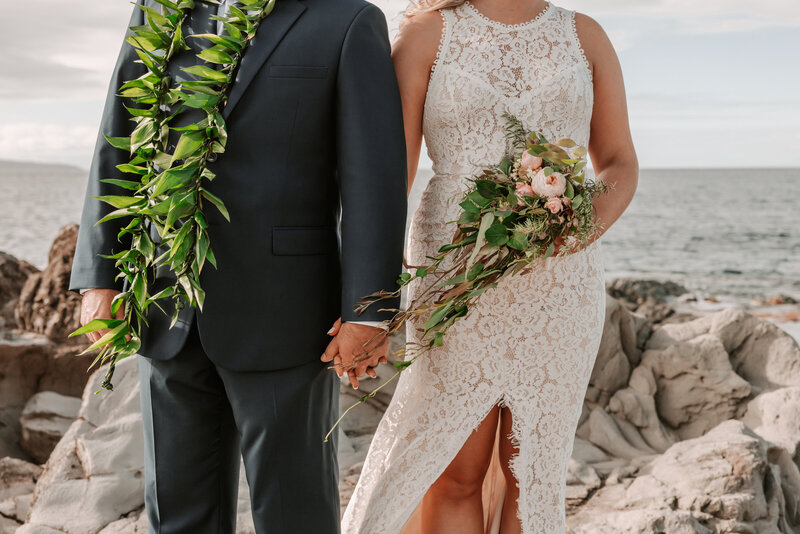 newly wed couple in Maui standing on a cliff edge after saying their personal vows. She is holding a bouquet and he has a green maile lei on. The wife is wearing a brides dress and he is wearing a tux