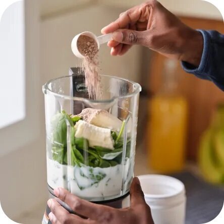 A stock photo of a hand pouring powdered supplement into a green smoothie, linking to Tracy Greenstein’s Fullscript premium supplement page