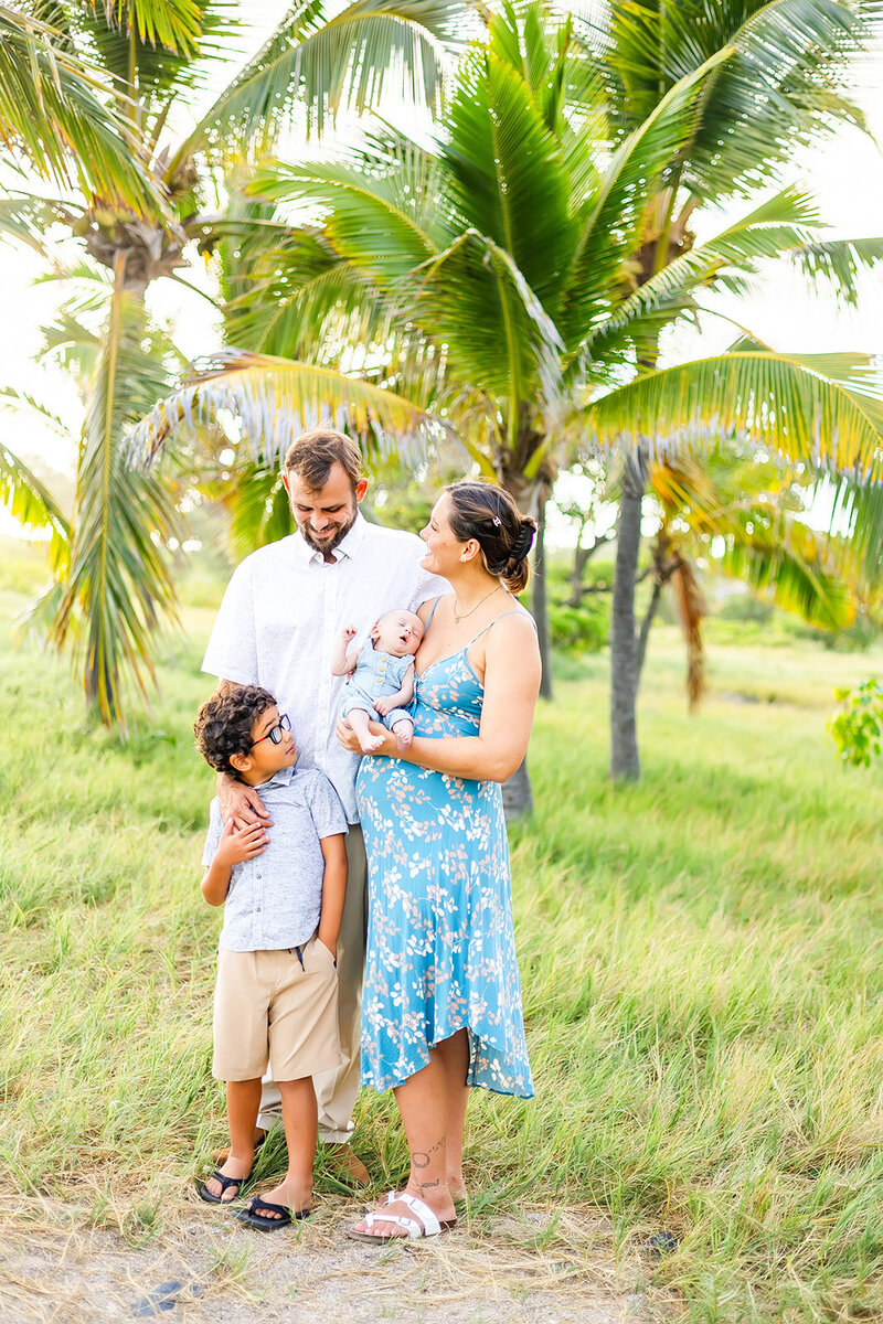 family with white shirt and blue dress stand  on a sandy beach laughing in front of palm trees