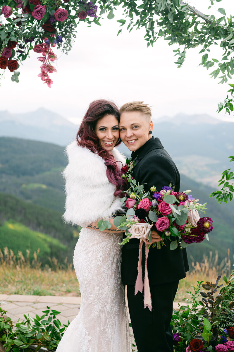Bride and bride walk hand in hand with hands up in the air in front of a flower arch and mountain background