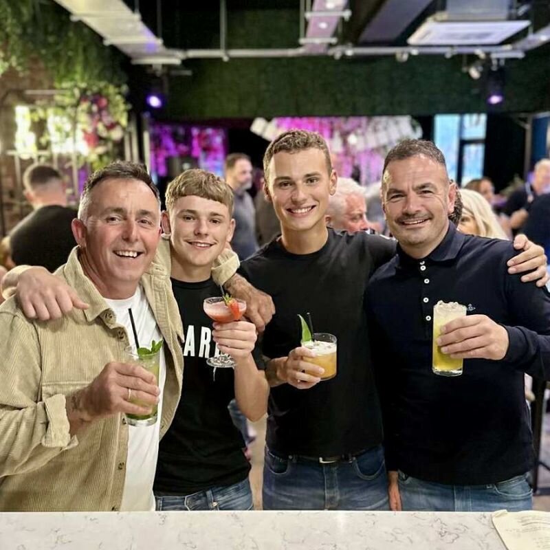 Four men posing with smiles at a bar with cocktails in hand.