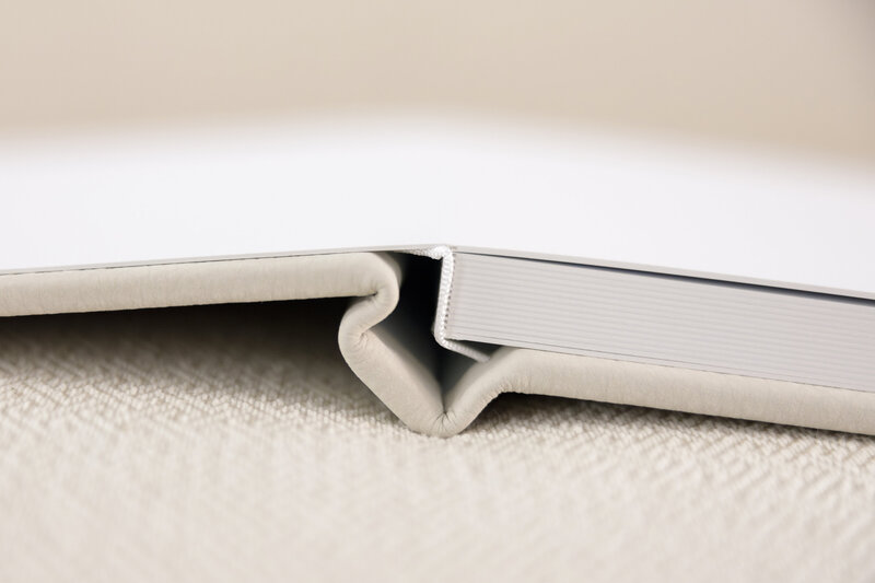 Close-up of an open hardcover album lying on a textured surface. The book is slightly ajar, revealing the edge of its white pages and a beige cover. The background is softly blurred, emphasizing the books details.