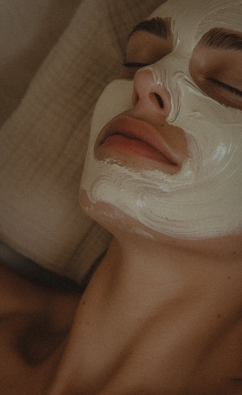 Close-up of a person indulging in a soothing facial treatment, lying down with a white mask applied, eyes closed, and lips slightly parted. The serene ambiance envelops them in tranquility.