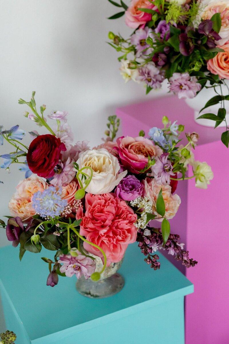 Colorful floral arrangement with roses, ranunculus, and garden blooms styled against pink and teal blocks by Tamara Menges Designs.