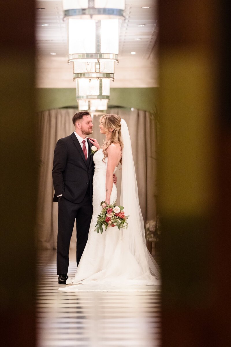 Bride and groom smiling at each other in an elegant hallway, framed between doorways, with soft chandelier light and a romantic atmosphere.