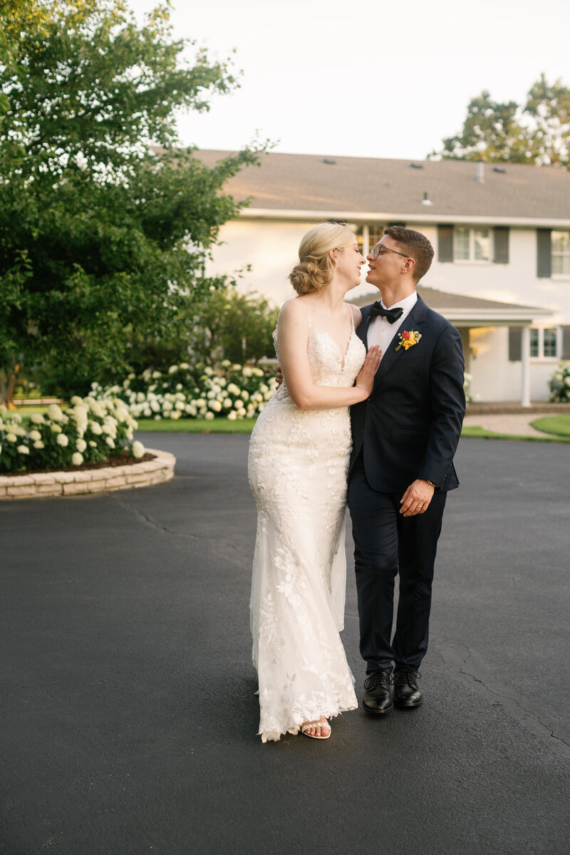 A wedding couple embraces romantically on a paved driveway with white flowering bushes in the background.