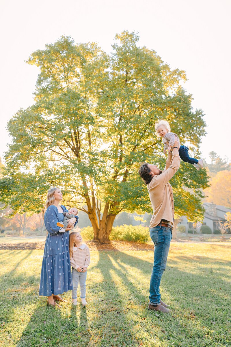 Family of five plays outside in front of big tree with sunlight streaming through during family photo session in Hendersonville, North Carolina with Rachel Marie Photography