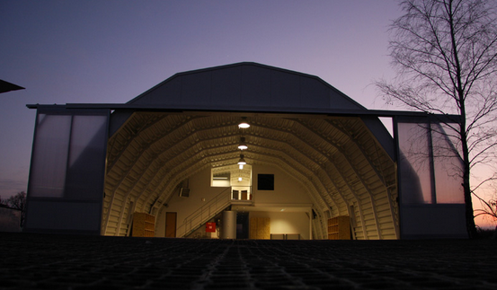 Der ATTC Hangar in Abendstimmung. Das Bild zeigt einen Hangar, der innen beleuchtet ist und mit geöffneten Türen in Abendstimmung