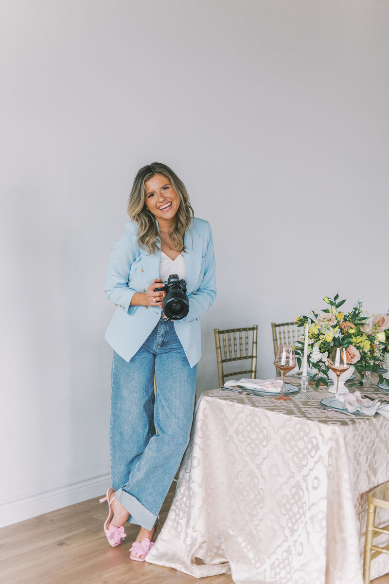 Headshot of Lindsey Kristian in a blue blazer and jeans holding her camera and standing beside a decorated tablescape