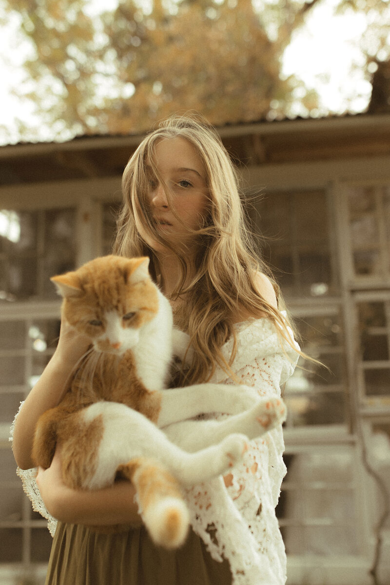 A senior girl with flowy blonde hair looks at the camera with a serious gaze while holding her orange cat. 